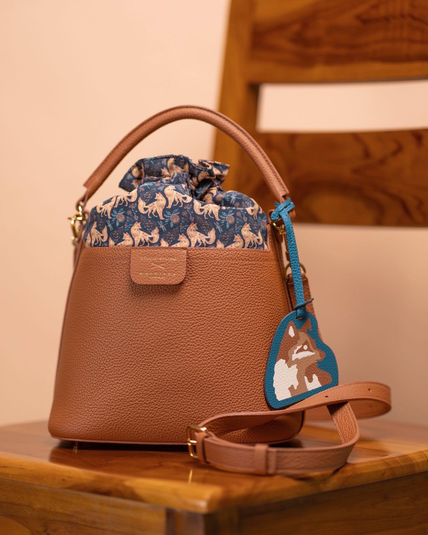 Teal Kitsune bag charm clipped to a tan leather bucket bag on a wooden chair