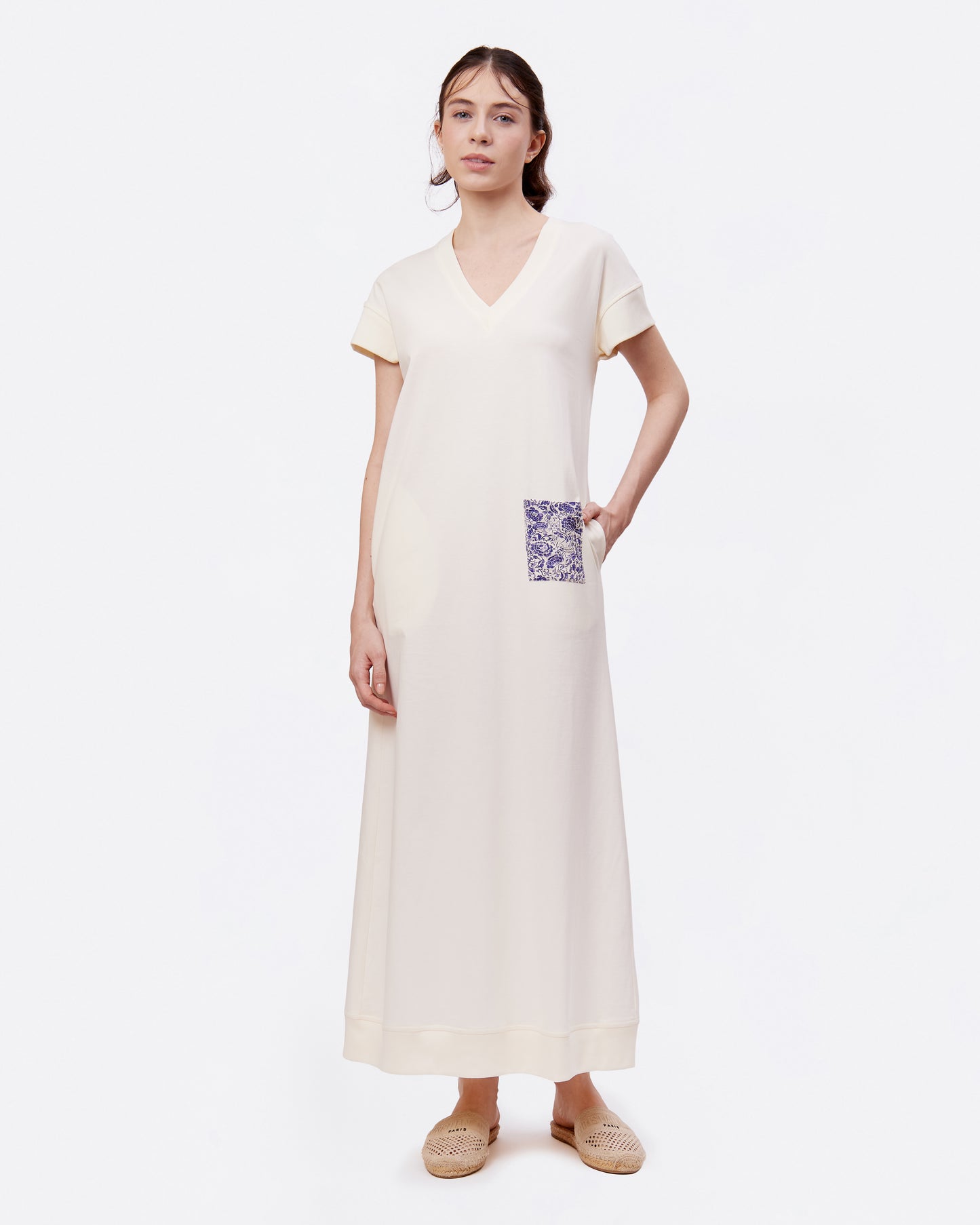 Woman wearing a long white cotton maxi dress with a printed pocket detail on a plain background