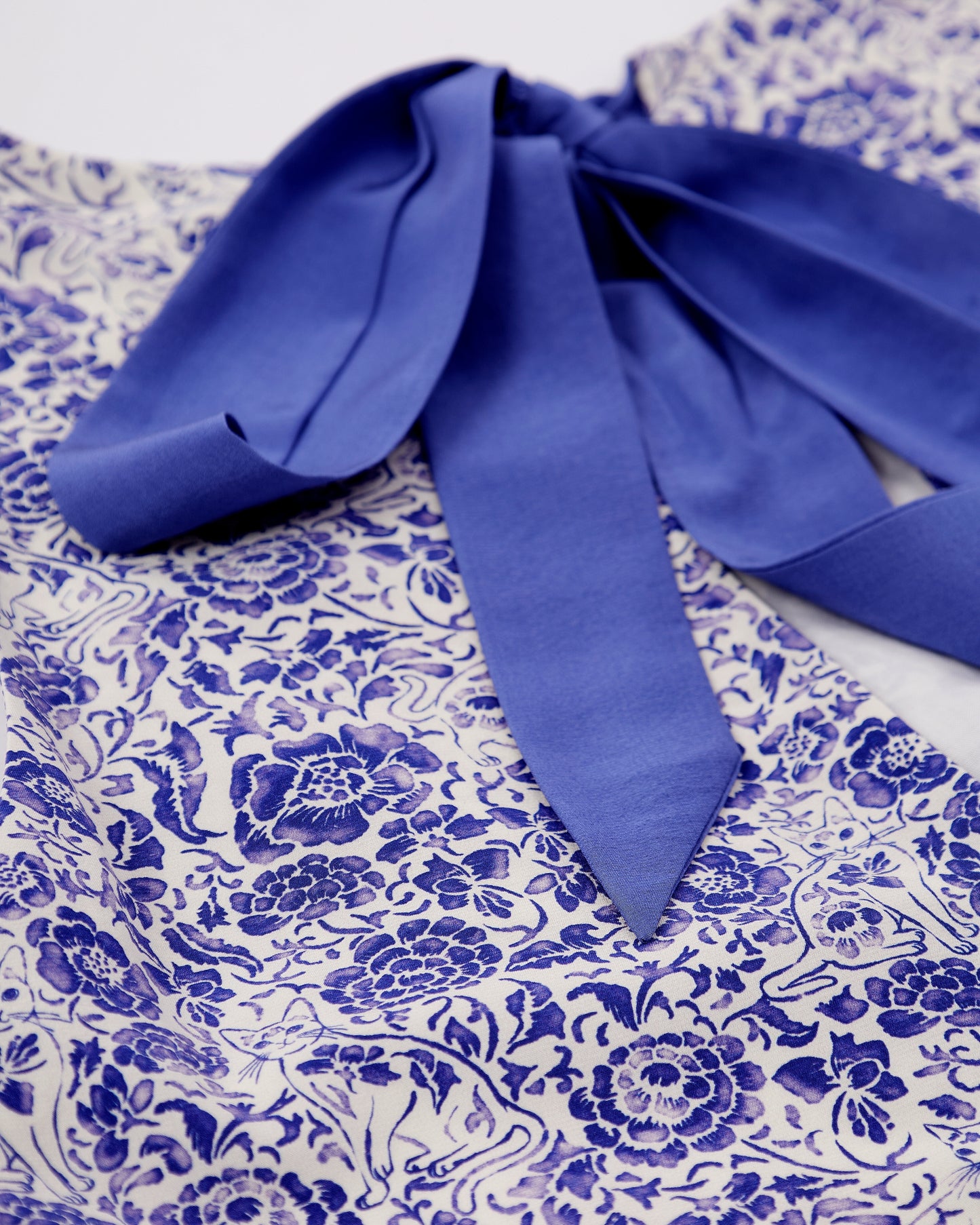 Close up of a blue floral and porcelain cat patterned silk blend dress with a ribbon detail on a white background