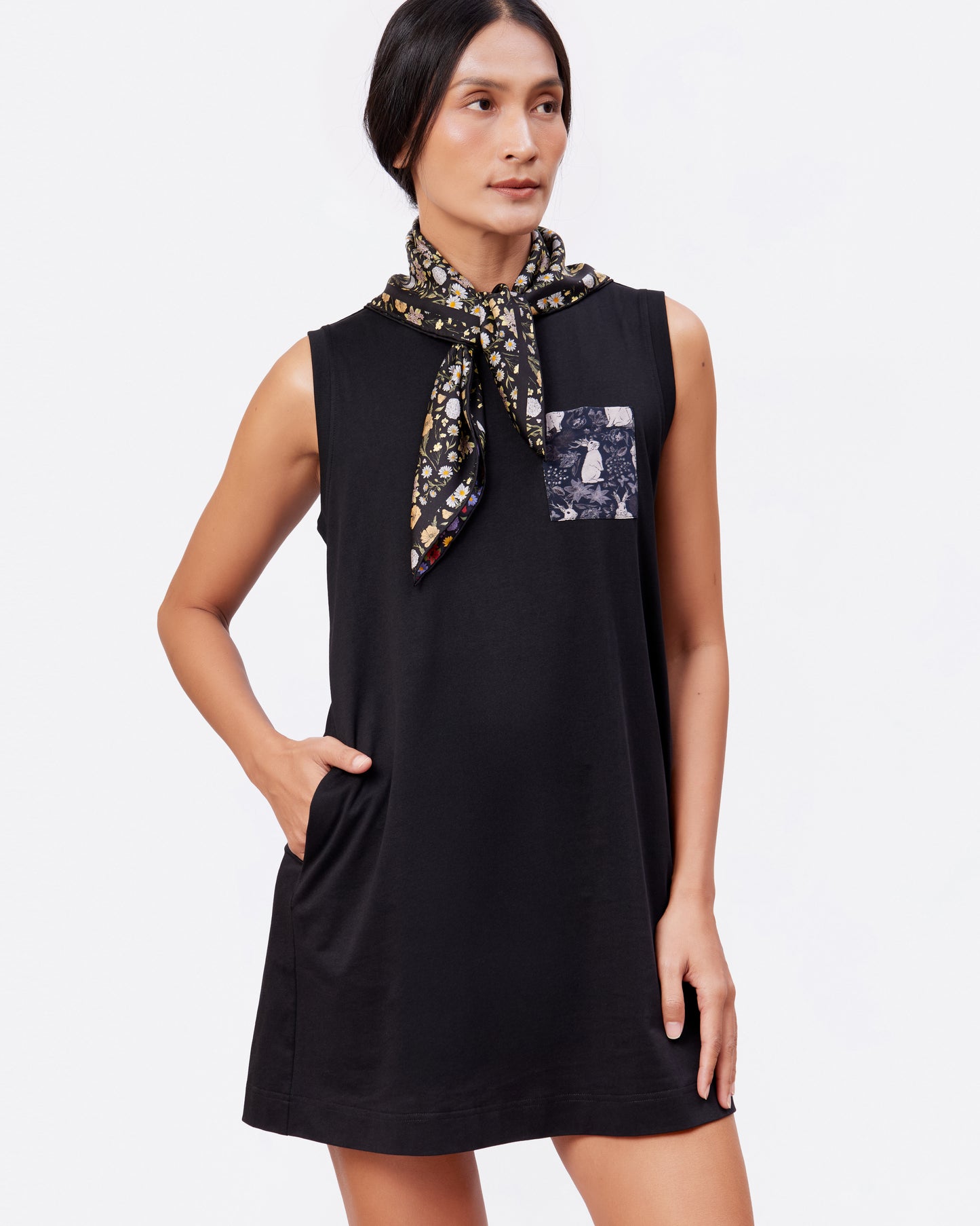Woman wearing a black sleeveless cotton tank dress with a printed pocket and a floral silk scarf on a white background