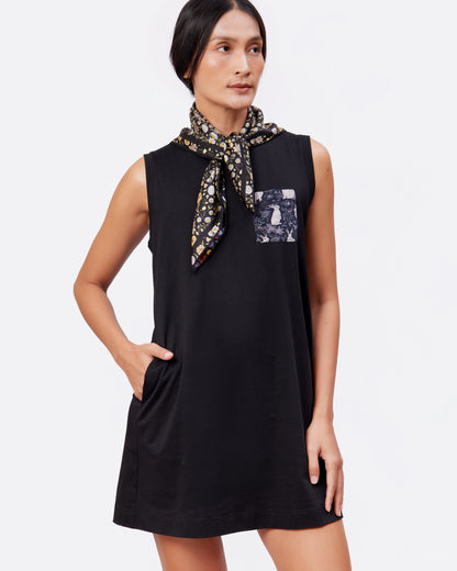Woman wearing a black sleeveless cotton tank dress with a printed pocket and a floral silk scarf on a white background