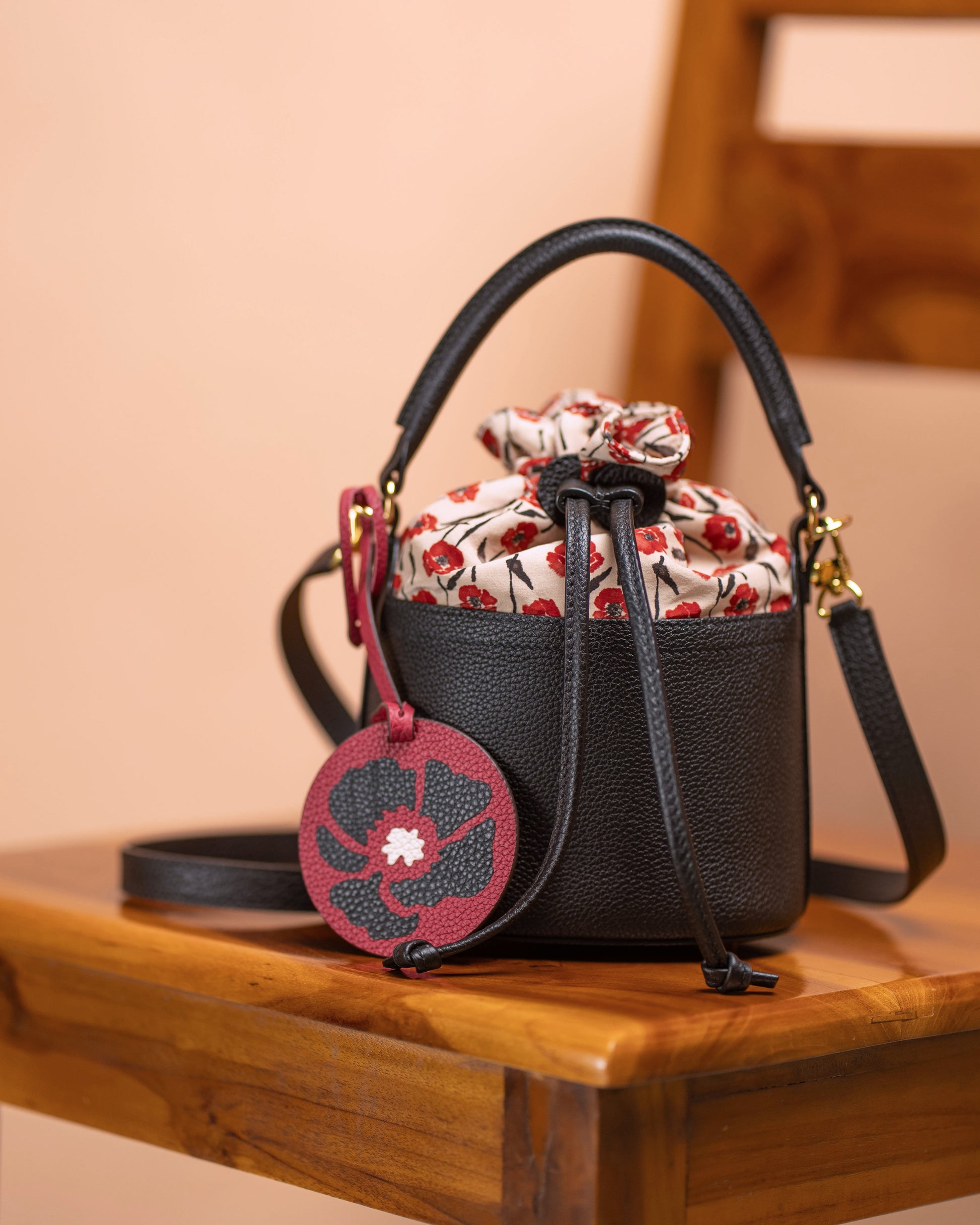 Red poppy bag charm attached to a black leather mini bucket bag on a wooden chair