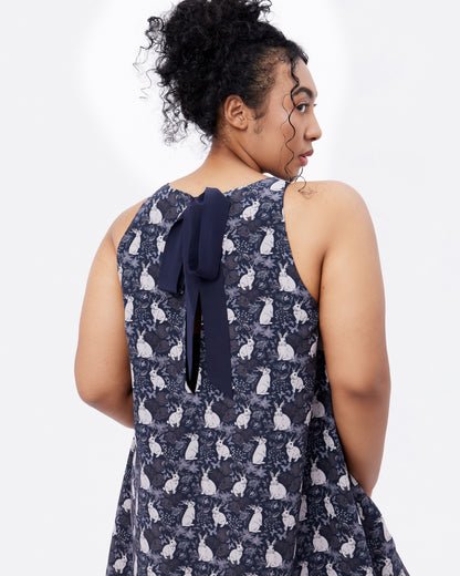 Woman wearing a black sleeveless silk blend dress with mythical rabbit pattern and bow detail on a plain background