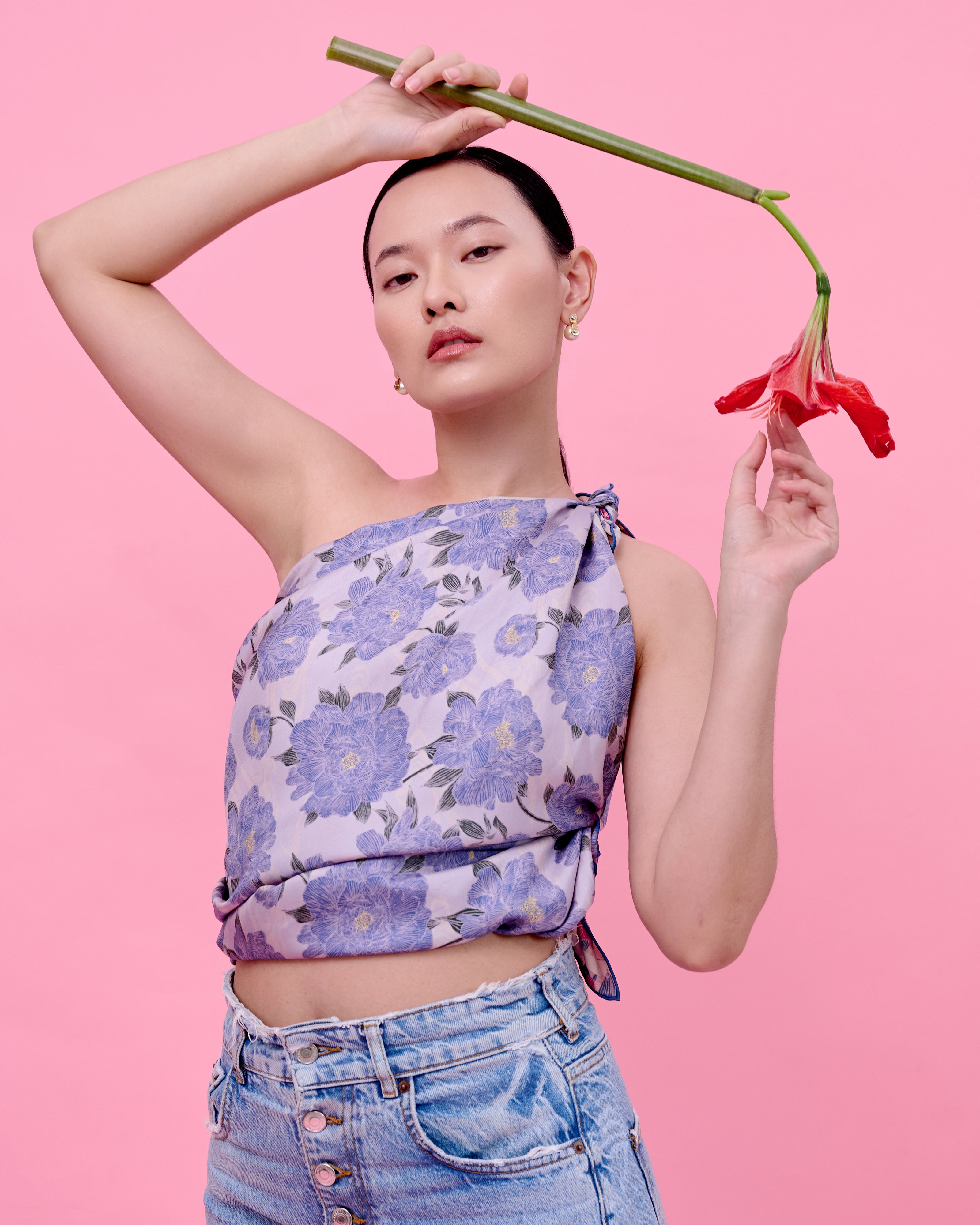 Woman wearing a floral silk scarf top and jeans holding a red flower against a pink background